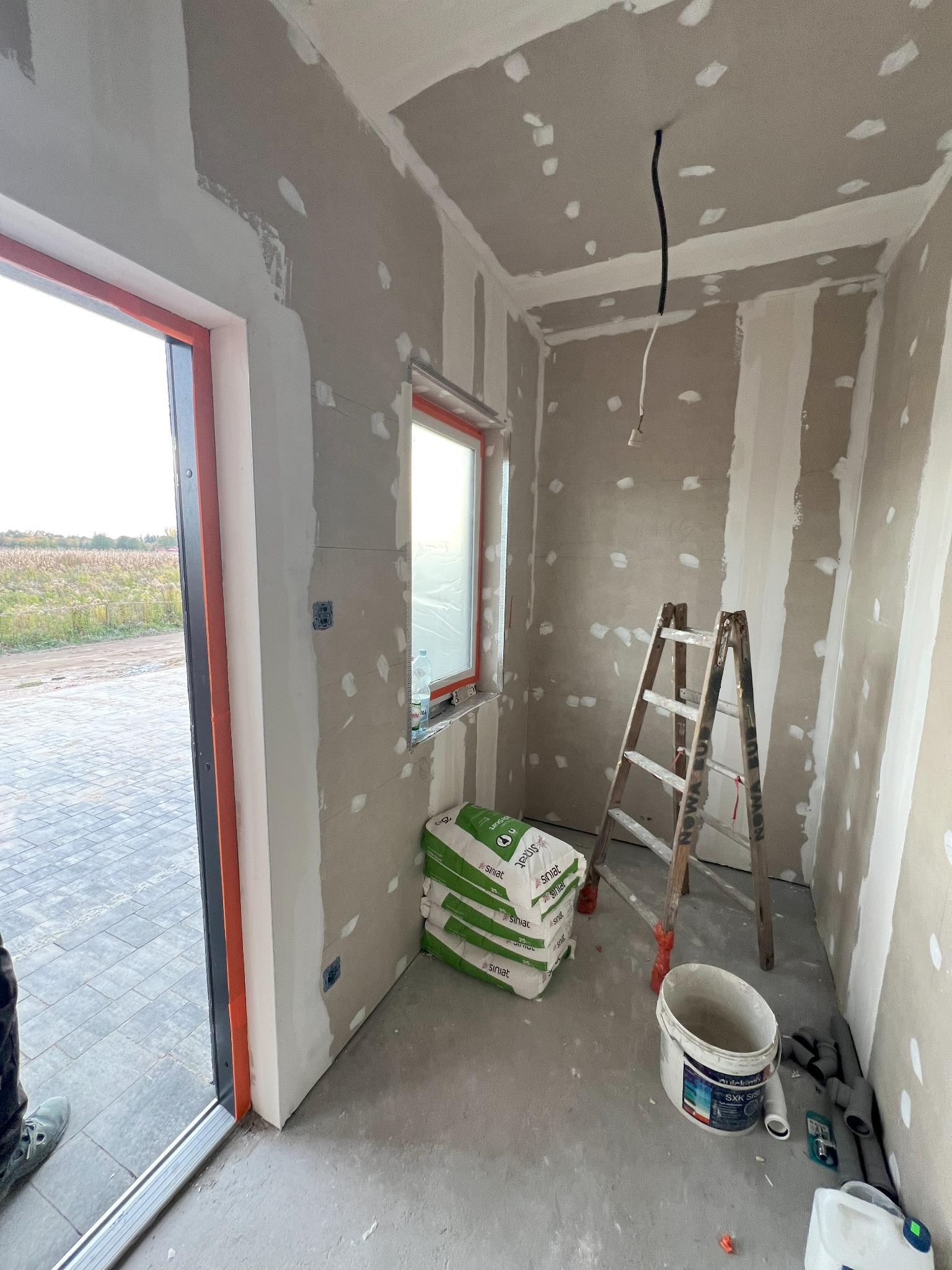 Wnętrze w trakcie prac wykończeniowych: ściany przygotowane do szpachlowania, widoczne łączenia płyt, drabina oparta o ścianę, worki z gładzią, wiadro z zaprawą i elementy instalacji kanalizacyjnej...