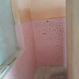 Raf bud - Ściana pomalowana na różowo z widocznymi plamami i niedoskonałościami, surowy stan przed wykończeniem, fragment podłogi i ściany w rogu pomieszczenia, widoczne ślady po szpachlowaniu.