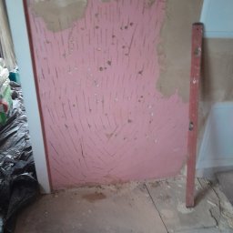 Raf bud - Ściana przygotowana pod szpachlowanie z widocznymi śladami po zdzieraniu starej farby, zabezpieczona różowym podkładem, obok pionowa poziomica i wystające kable ze ściany.