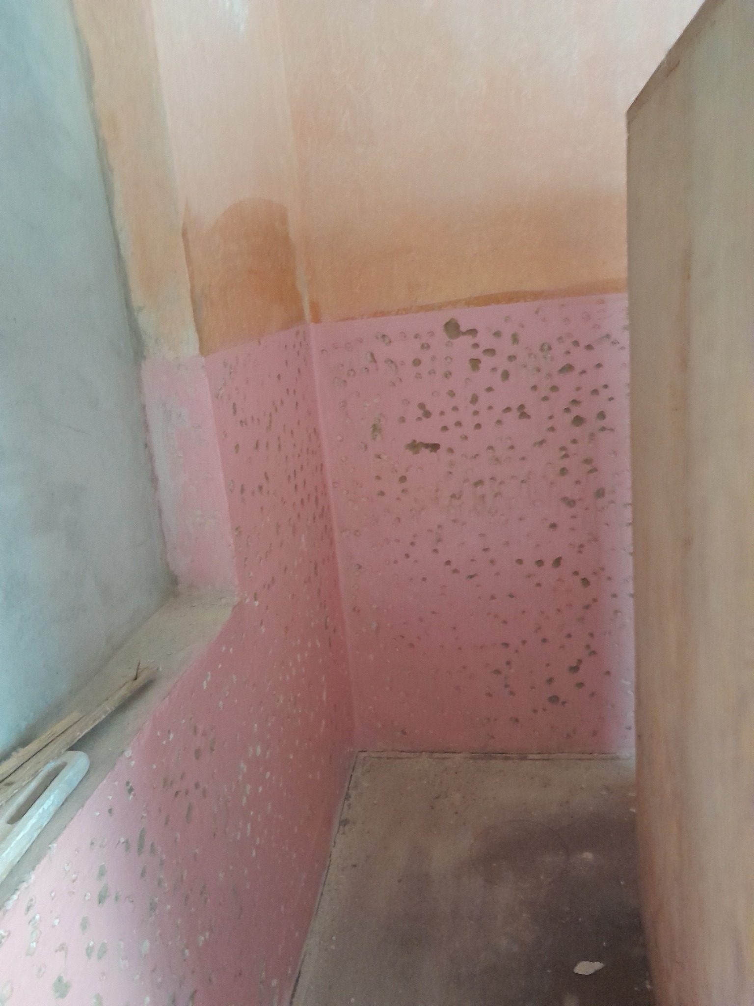 Ściana pomalowana na różowo z widocznymi plamami i niedoskonałościami, surowy stan przed wykończeniem, fragment podłogi i ściany w rogu pomieszczenia, widoczne ślady po szpachlowaniu.