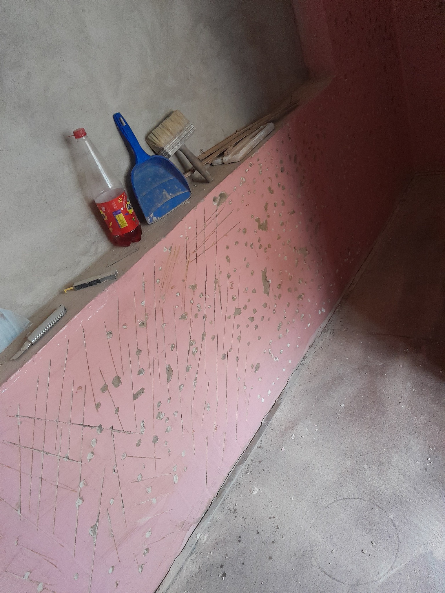 Ściana przygotowana do dalszych prac wykończeniowych, z widocznymi śladami po szpachlowaniu i rysami, obok leżą narzędzia: szpachelka, pędzel, nożyki oraz butelka z napojem.