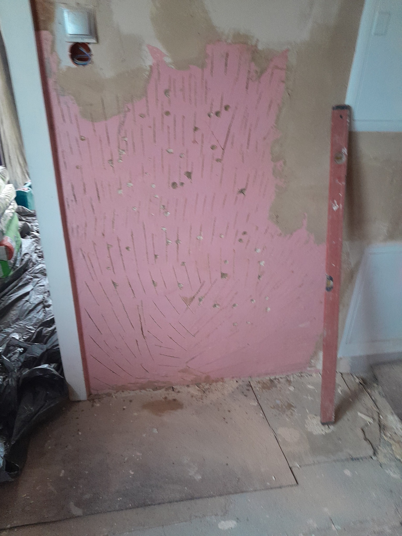 Ściana przygotowana pod szpachlowanie z widocznymi śladami po zdzieraniu starej farby, zabezpieczona różowym podkładem, obok pionowa poziomica i wystające kable ze ściany.