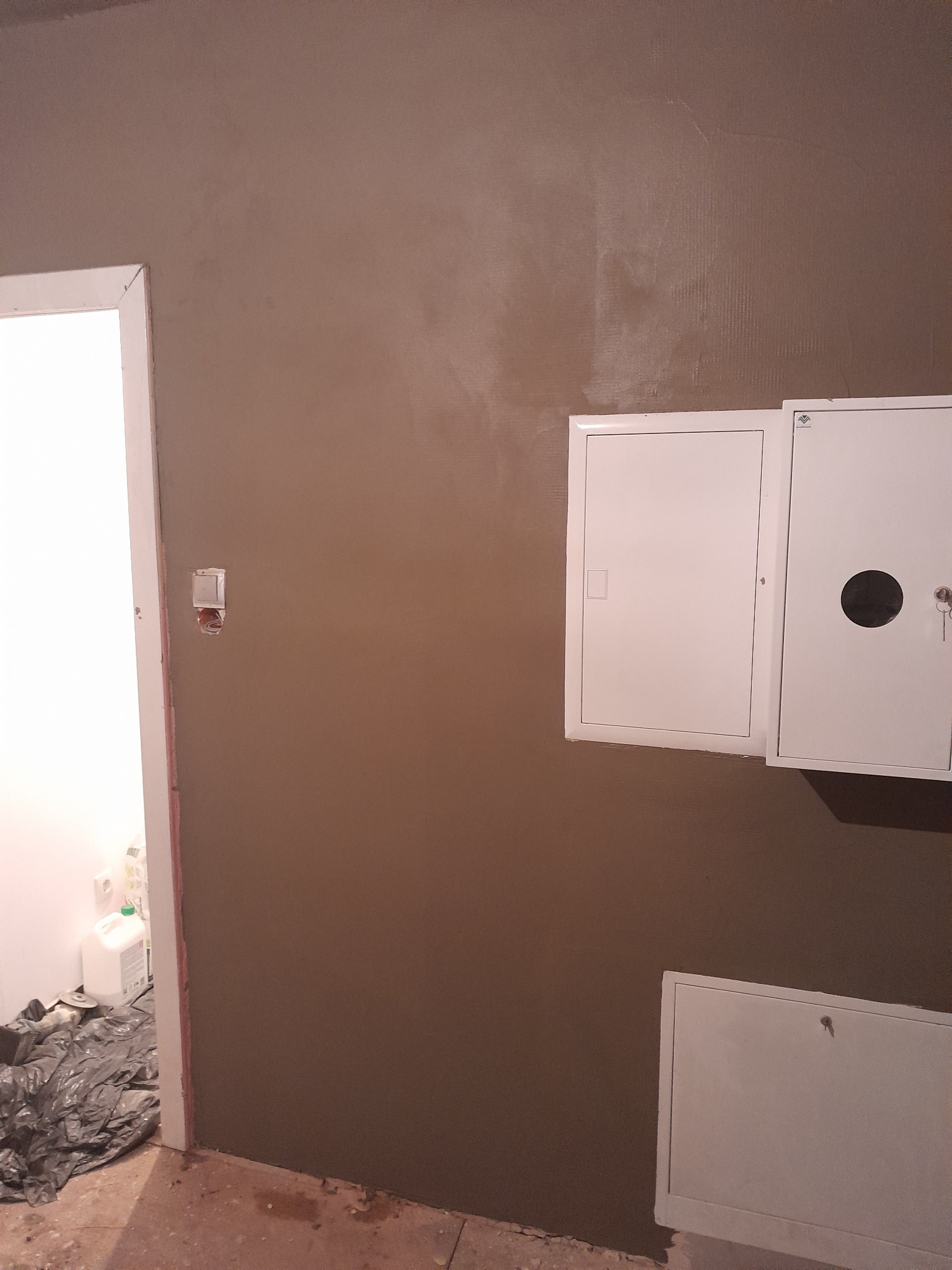 Ściana po nałożeniu gładzi szpachlowej, widoczne białe skrzynki elektryczne, obok ościeżnicy widoczne wiadro z płynem i folia ochronna.