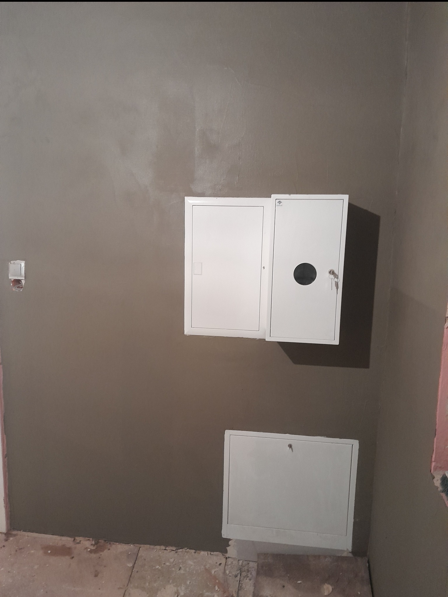 Świeżo nałożona, szara gładź na ścianie z zamontowanymi białymi skrzynkami instalacyjnymi; widoczne ślady po pracach na podłodze i niedokończone wykończenie przy futrynie.