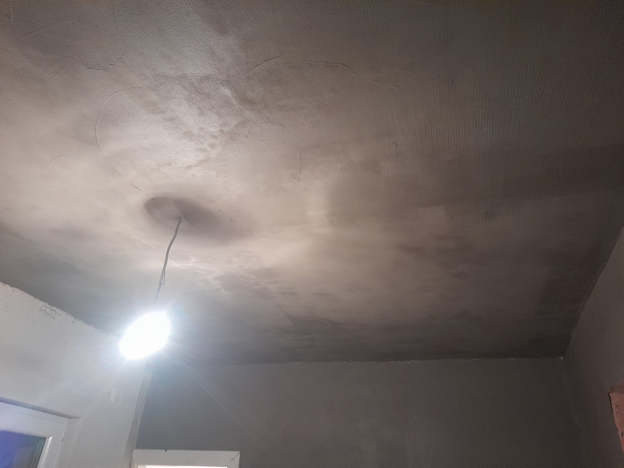 Niedokończony sufit po nałożeniu warstwy szpachli, widoczna wisząca żarówka i fragment ściany w szarym odcieniu.