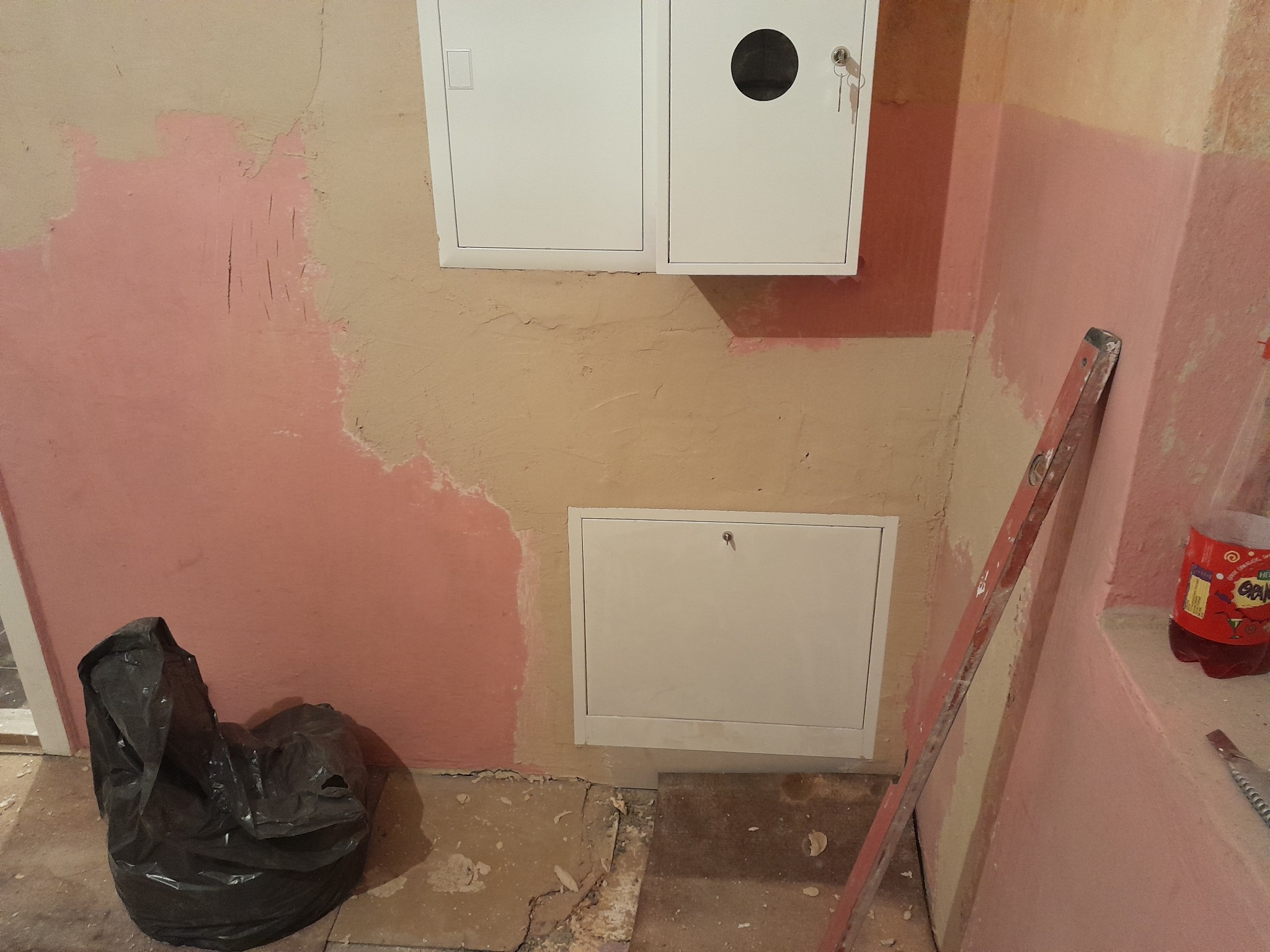 Ściana w trakcie przygotowania do prac wykończeniowych, widoczne fragmenty starej farby, dwie białe skrzynki rewizyjne, poziomica oparta o ścianę, worek na śmieci i butelka napoju.