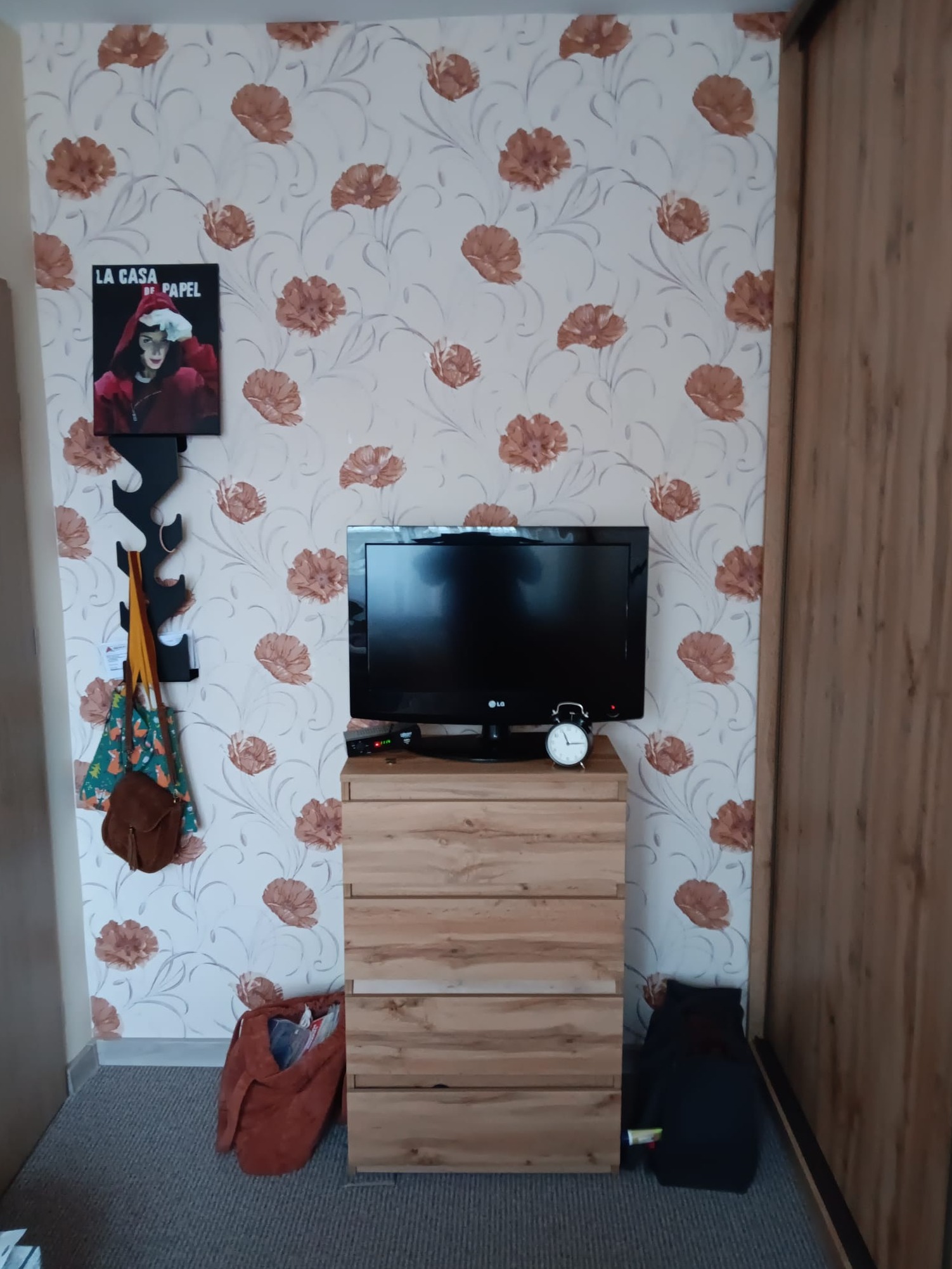 Pokój z tapetą w kwiaty, komodą z telewizorem i dekoracjami ściennymi, w tym plakat La Casa de Papel i wieszak na ubrania.