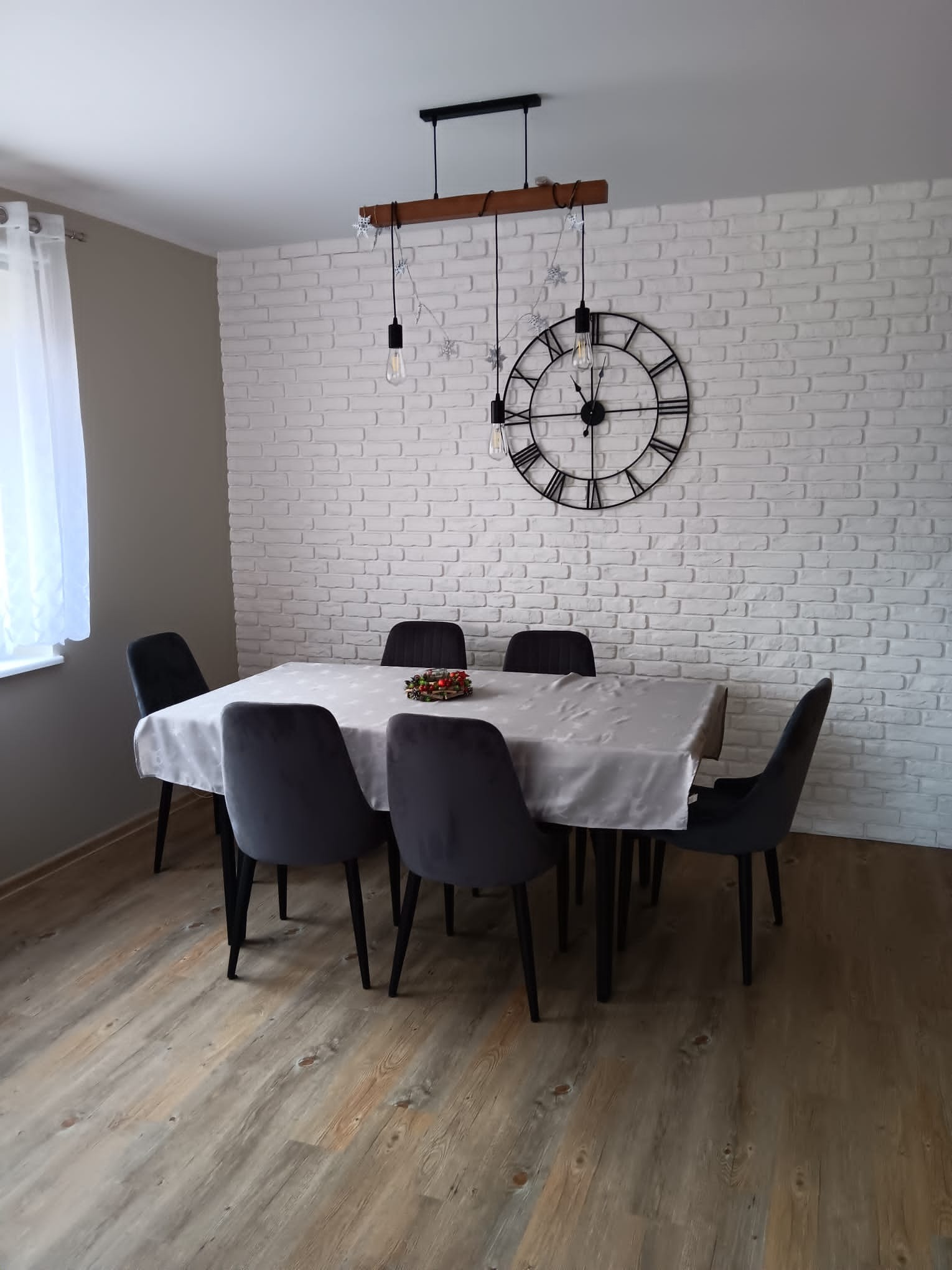 Jadalnia z białej cegły z drewnianą lampą wiszącą, zegarem ściennym i szarymi krzesłami welurowymi wokół stołu przykrytego jasnym obrusem z subtelnym wzorem.