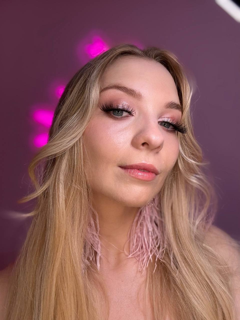 Portret blondynki z delikatnym, różowym cieniowaniem powiek, wydłużonymi rzęsami i błyszczącymi ustami, uzupełniony kolczykami z piór w odcieniu pudrowego różu na tle fioletowej ściany z efektem...