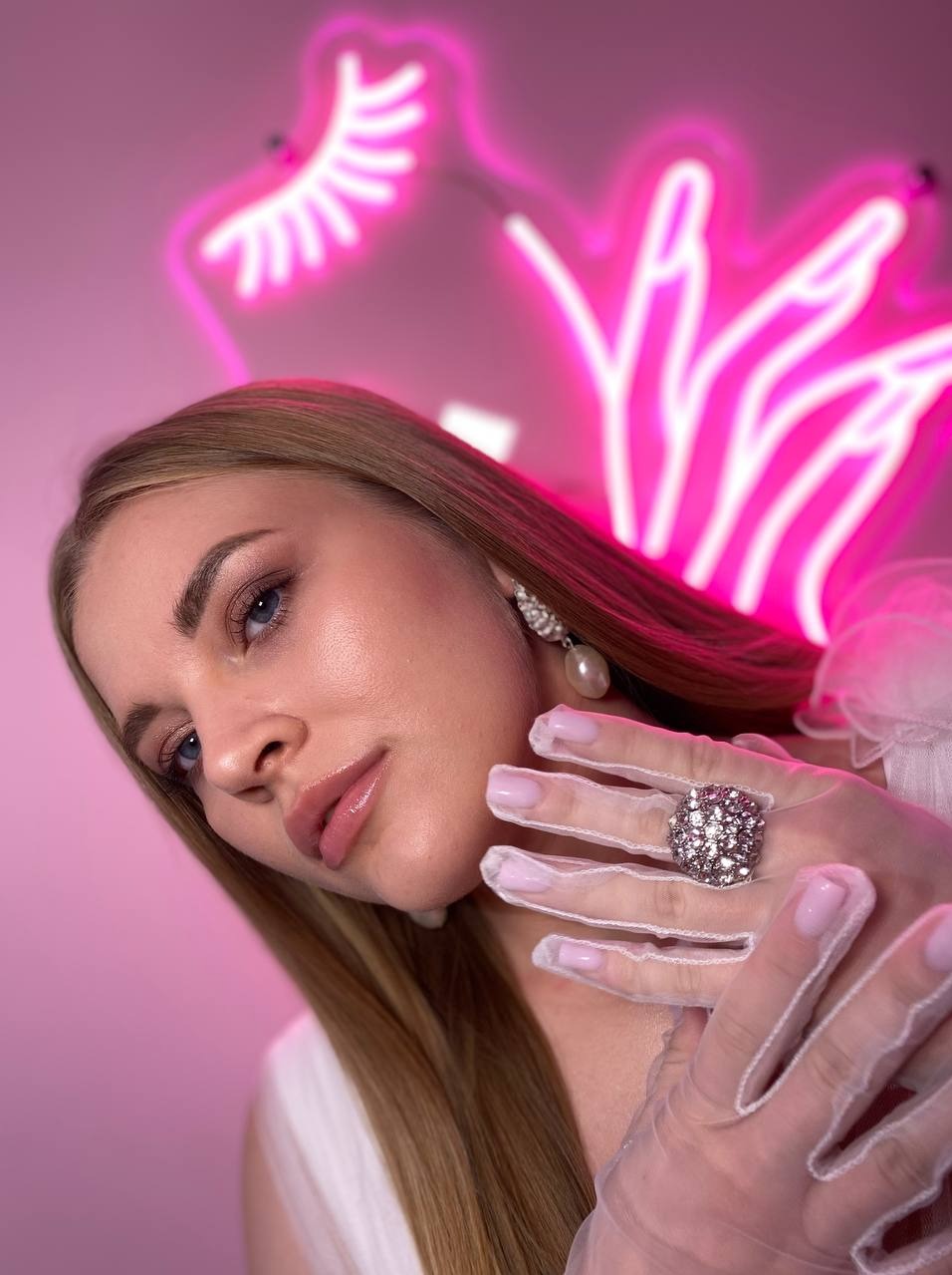 Portret kobiety z delikatnym makijażem oczu, perłową biżuterią i dużym pierścieniem na tle neonowego znaku w kształcie rzęs.