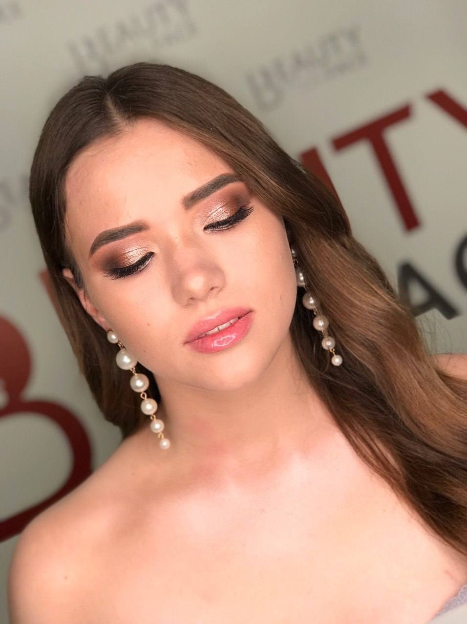 Portret kobiety z profesjonalnym, błyszczącym makijażem oczu w odcieniach brązu i różu, pełnymi ustami z błyszczykiem oraz długimi perłowymi kolczykami na tle logo salonu.