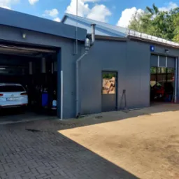 Zewnętrzny widok warsztatu samochodowego z dwoma otwartymi bramami, w jednej biały samochód osobowy, w drugiej widoczne wyposażenie warsztatowe, na zewnątrz narzędzia i pojemniki z płynami...