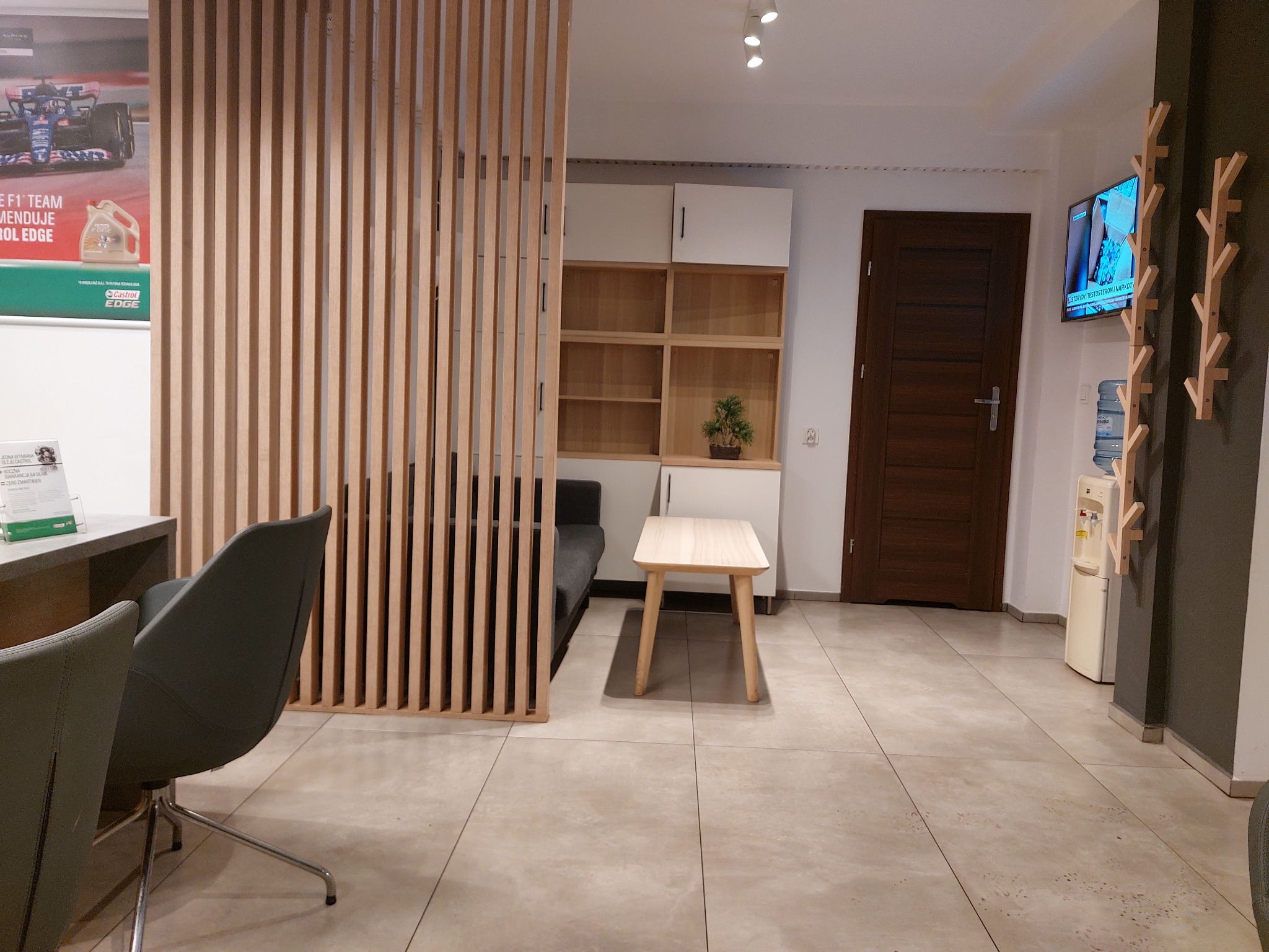 Poczekalnia warsztatu samochodowego z drewnianą ścianką działową, kanapą, stolikiem, telewizorem i dystrybutorem wody. Na ścianie plakat reklamowy oleju Castrol EDGE.