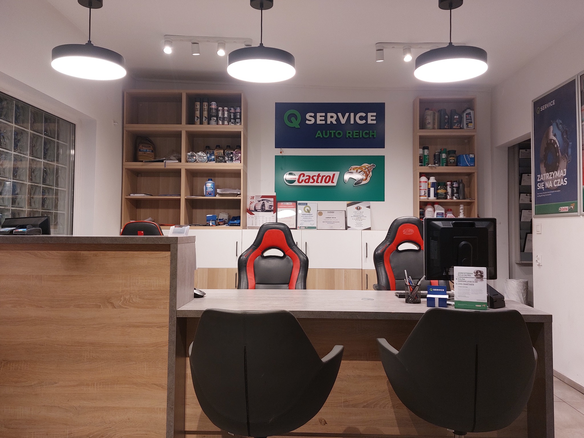 Recepcja warsztatu samochodowego z logo Q Service Auto Reich i Castrol, regały z olejami, krzesła biurowe w stylu sportowym.
