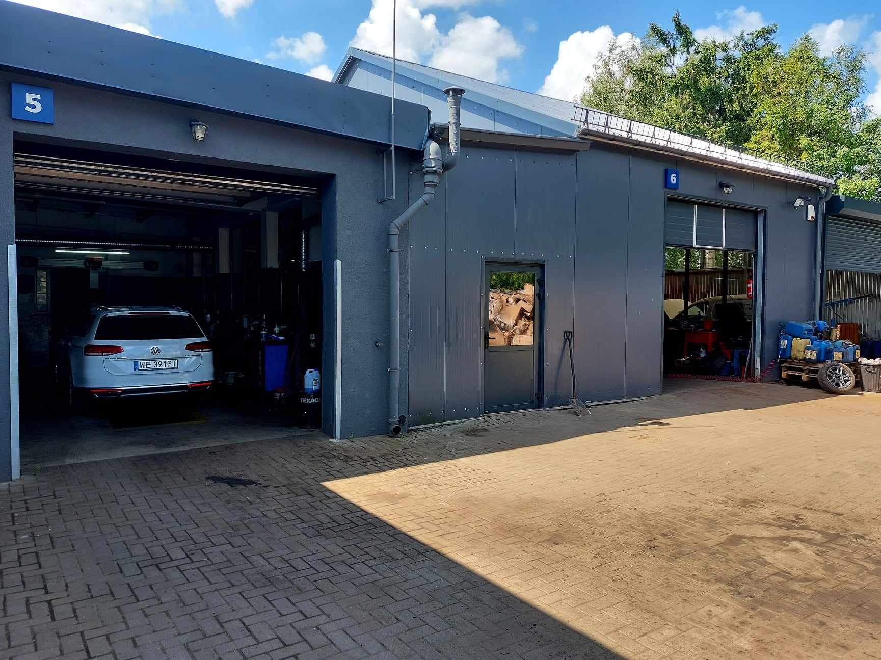 Zewnętrzny widok warsztatu samochodowego z dwoma otwartymi bramami, w jednej biały samochód osobowy, w drugiej widoczne wyposażenie warsztatowe, na zewnątrz narzędzia i pojemniki z płynami...