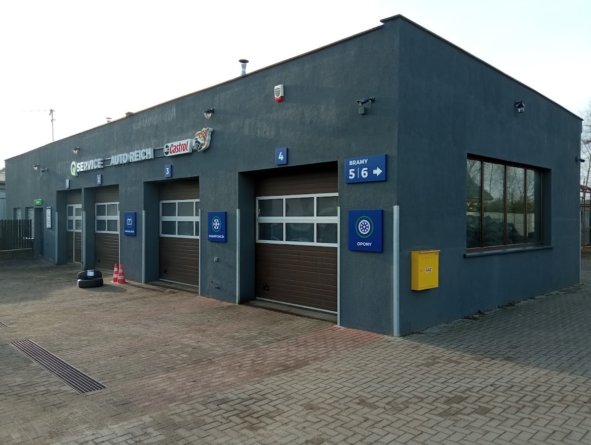 Zewnętrzny widok warsztatu samochodowego z trzema bramami wjazdowymi, oznaczeniami stanowisk i logo Castrol na elewacji. Na pierwszym planie opony i pachołki ostrzegawcze.