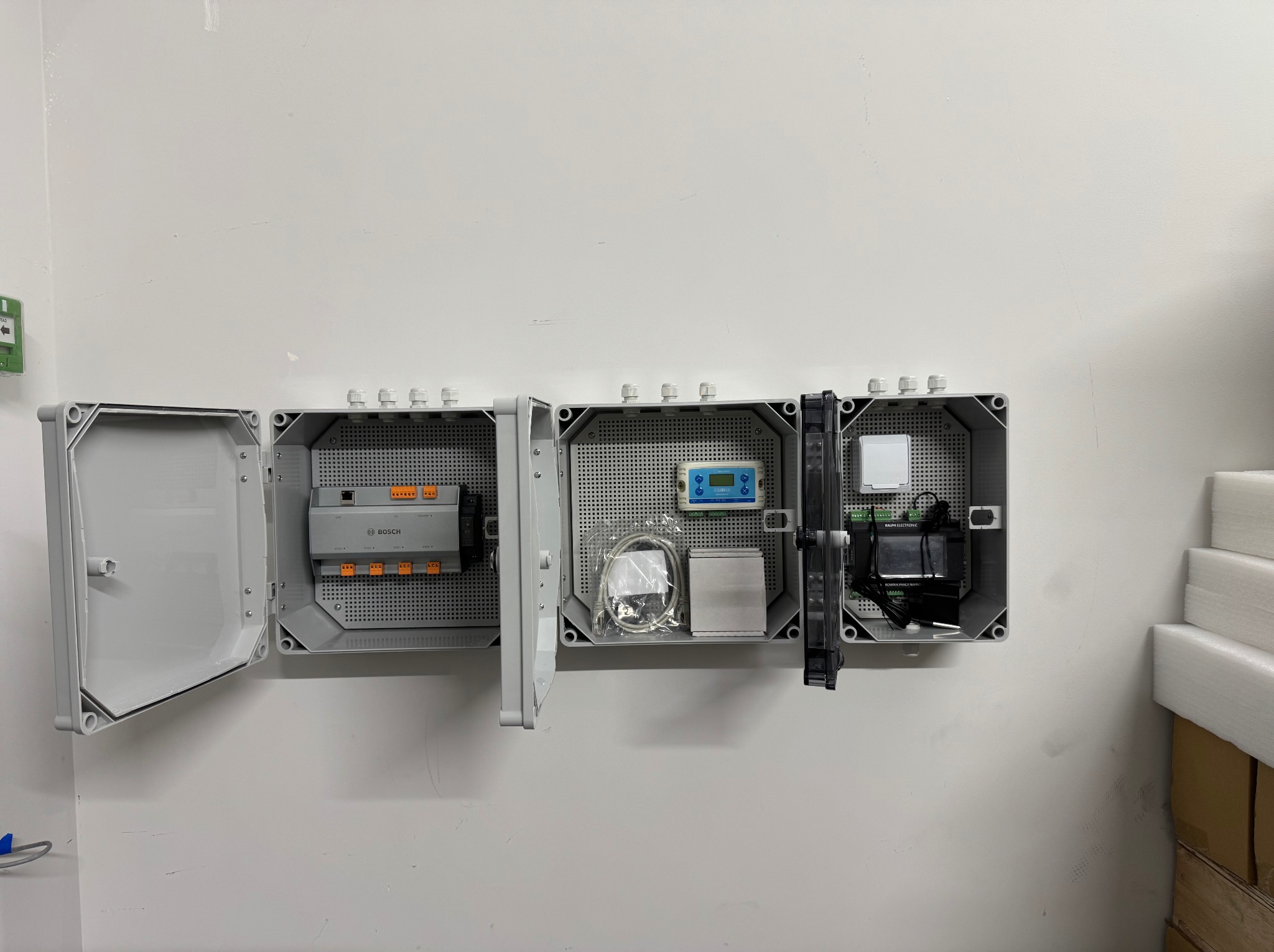 Otwarta, szara skrzynka elektryczna Bosch z modułami, okablowaniem i sterownikiem na białej ścianie. Widoczne elementy instalacji i białe piankowe płyty obok.