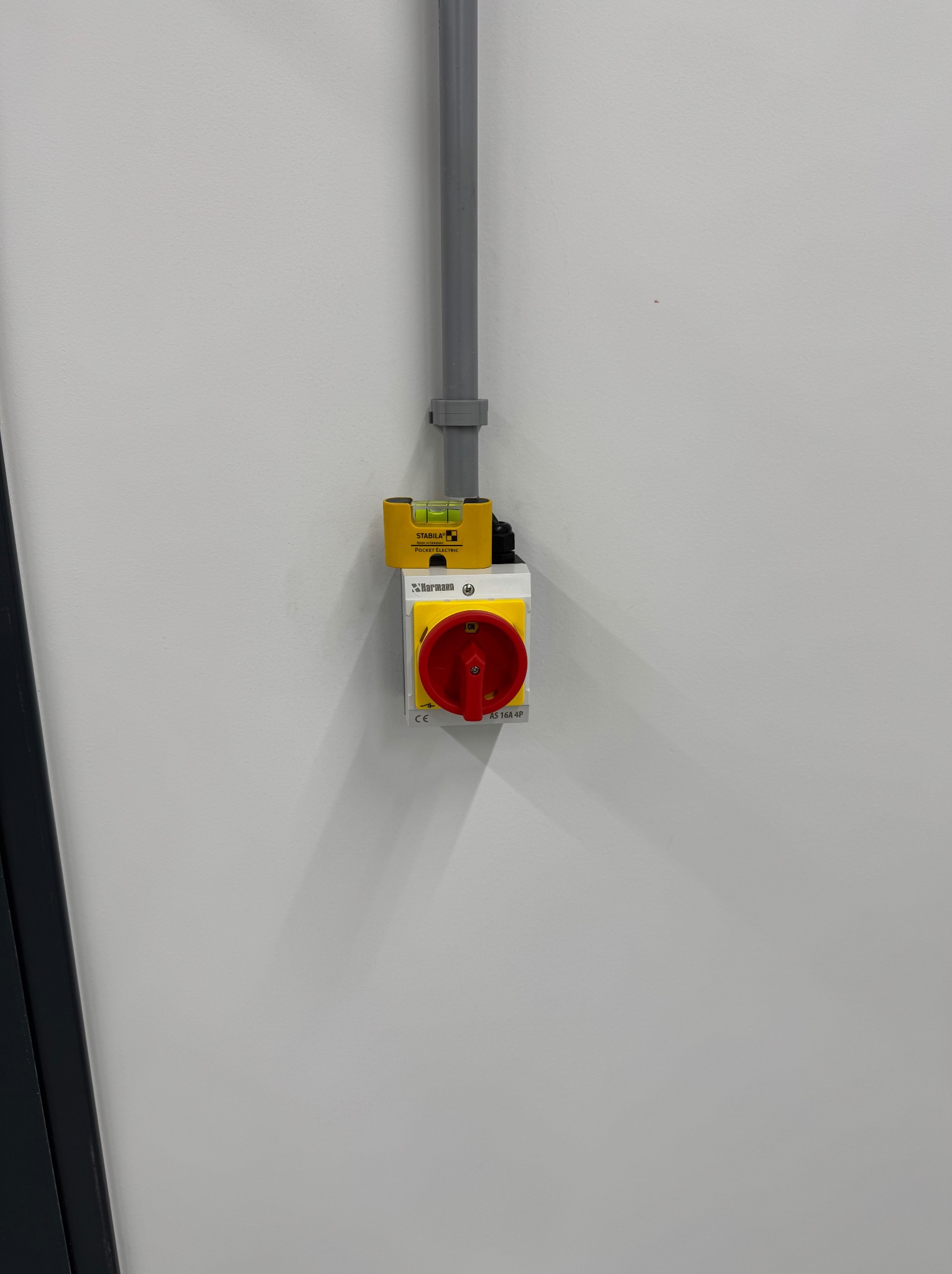 Zamontowany wyłącznik prądu z czerwoną dźwignią, przymocowany do ściany. Nad nim rurka instalacyjna i poziomica. Widoczne oznaczenia producenta i parametry techniczne.