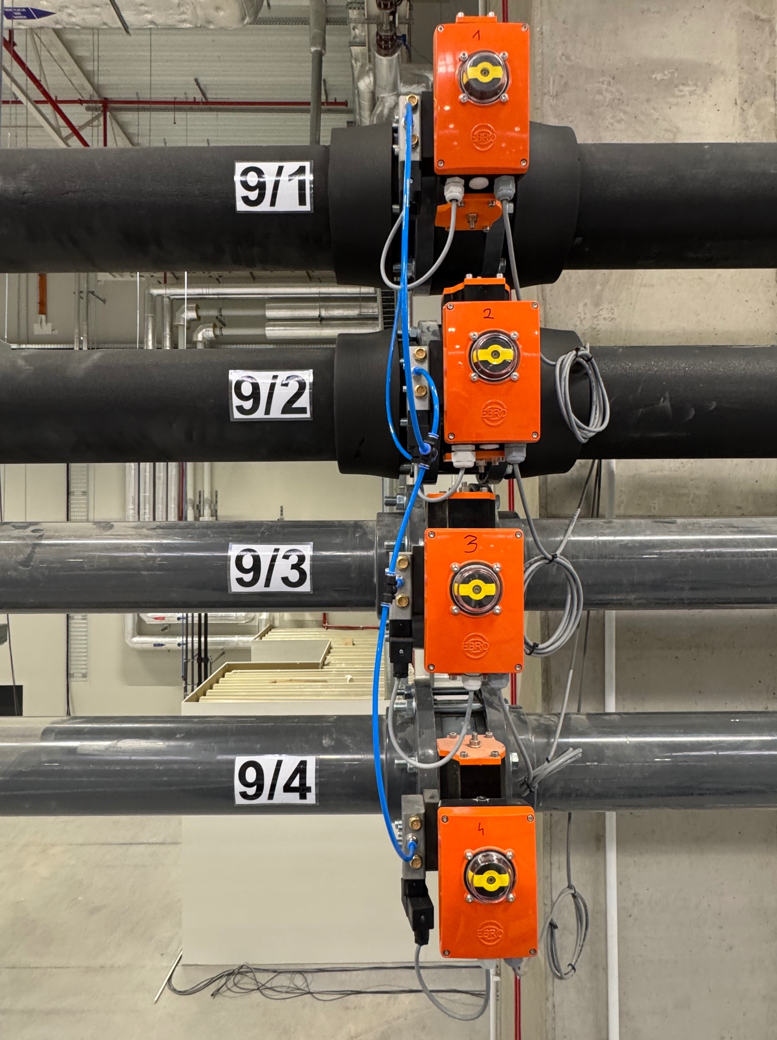 Cztery rury z czarną izolacją i pomarańczowymi, numerowanymi zaworami z żółtymi elementami sterującymi, zamontowane wzdłuż szarej ściany w budynku przemysłowym.
