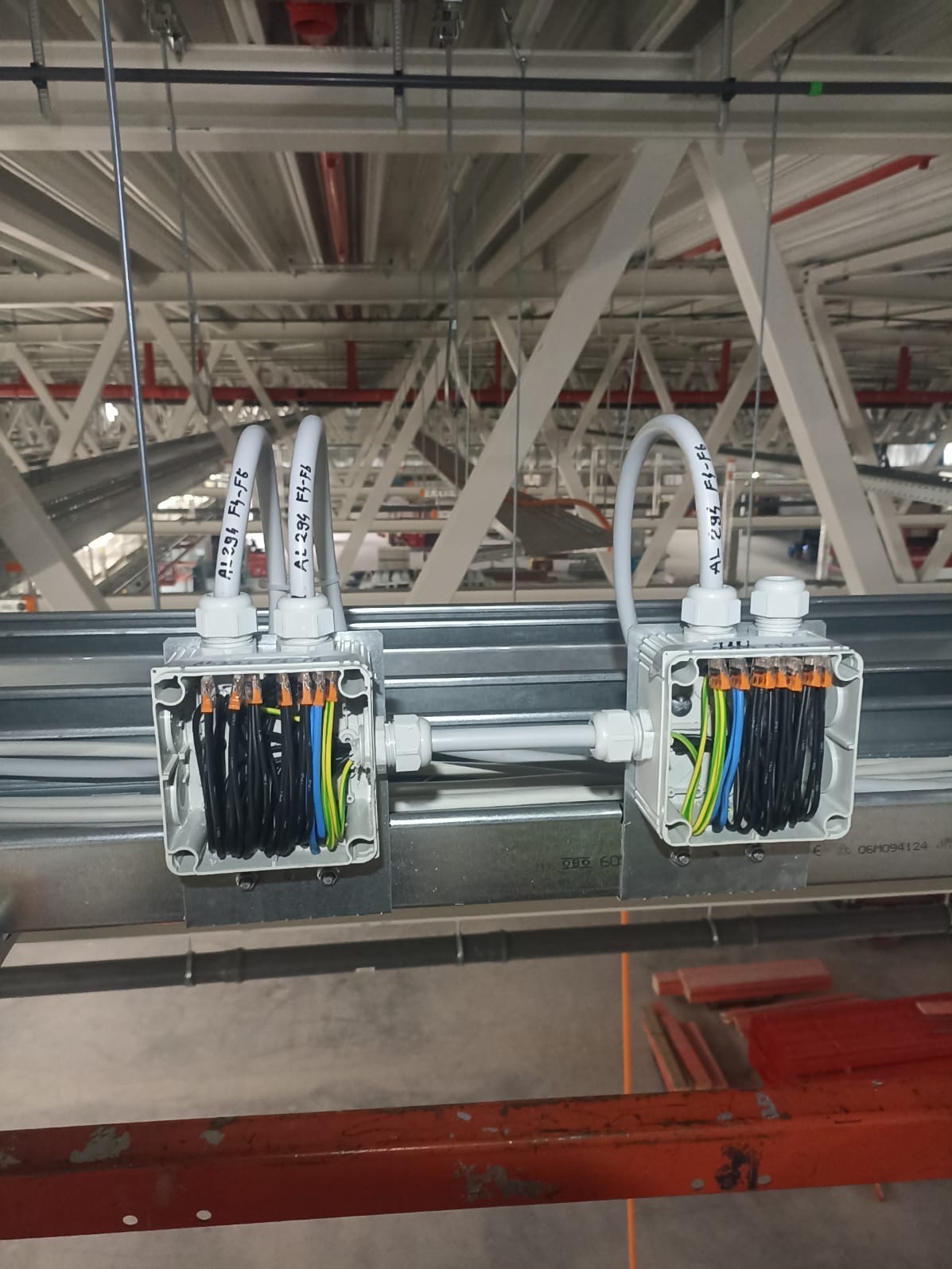 Dwa otwarte, szare puszki elektryczne z podłączonymi przewodami, zamontowane na metalowej konstrukcji pod sufitem w hali przemysłowej. Widoczne złącza WAGO i opis na peszlach.