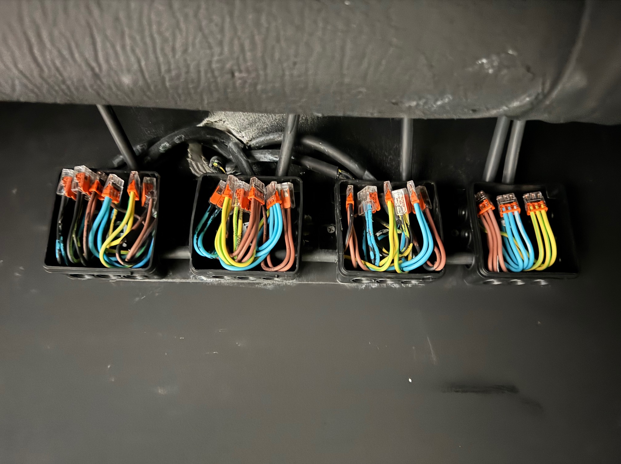 Cztery czarne puszki z kolorowymi przewodami (żółty, niebieski, brązowy) połączonymi złączkami Wago, zamontowane na szarej powierzchni. Widoczne kable w górnej części.