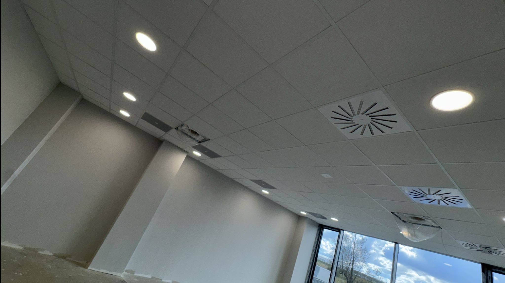 Biały sufit kasetonowy z zamontowanymi okrągłymi lampami LED i kratkami wentylacyjnymi. Widoczne okna i fragment ściany. Ujęcie z dołu, szeroki kąt.
