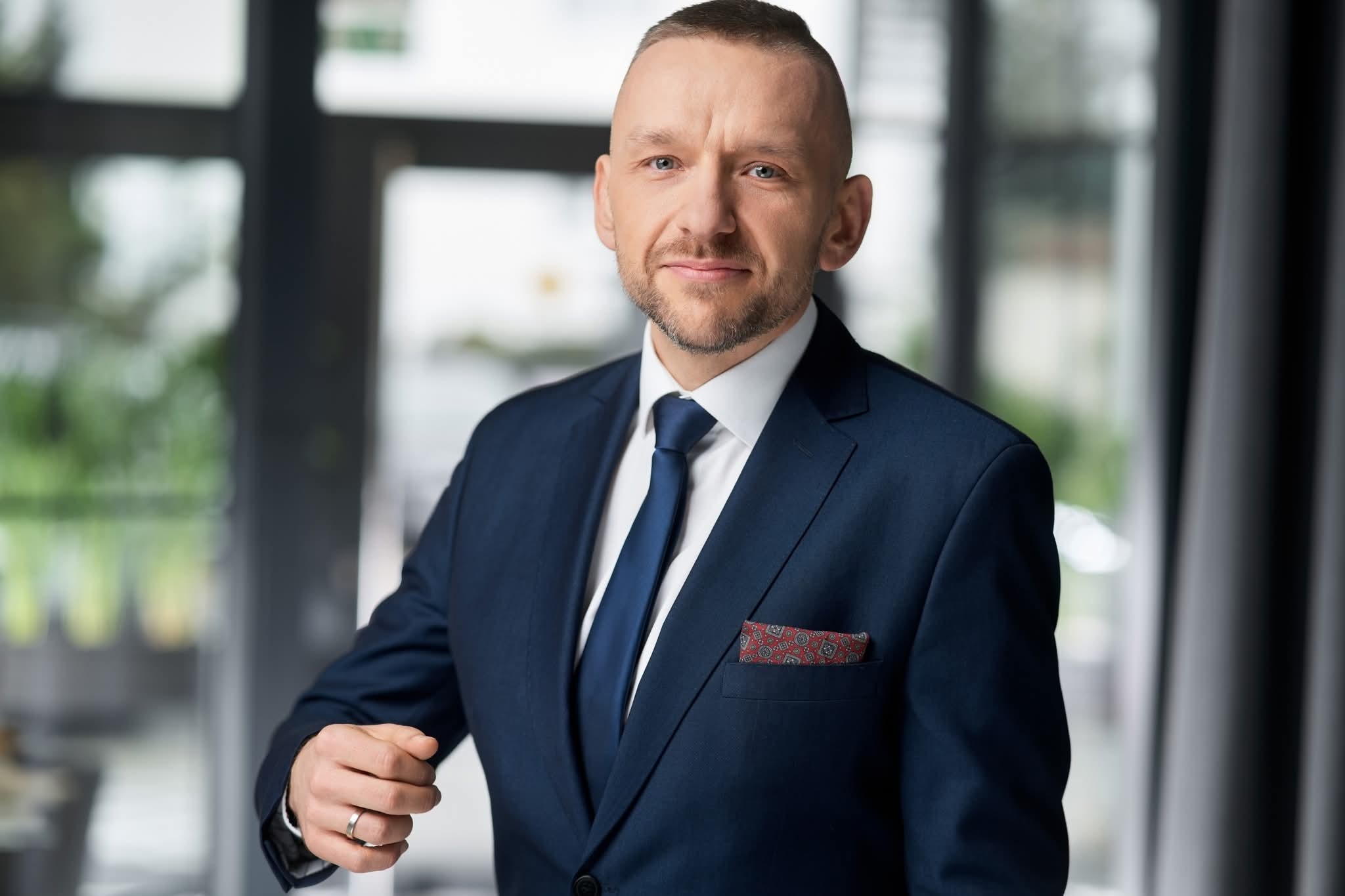 Portret mężczyzny w granatowym garniturze, białej koszuli i niebieskim krawacie. W tle okno z widokiem na zieleń. Widoczny pierścionek na palcu.