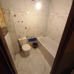 Łazienkowe Love - Przystępne cenowo i stylowo - Łazienka w trakcie remontu: białe płytki imitujące marmur na ścianach i podłodze, widoczne dystanse do płytek, przygotowane otwory na armaturę, biała wanna i toaleta, ślady prac remontowych.