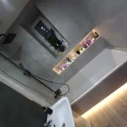 Nowoczesna łazienka z szarymi płytkami, czarną armaturą, podświetlaną wnęką na kosmetyki, oświetleniem pod wanną i rośliną w oknie.