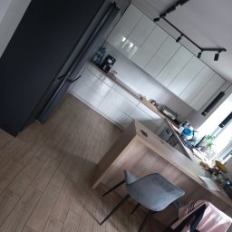 Łazienkowe Love - Przystępne cenowo i stylowo - Nowoczesna kuchnia z białymi szafkami na wysoki połysk, drewnianym blatem, podłogą z paneli imitujących drewno i czarnymi akcentami oświetleniowymi.