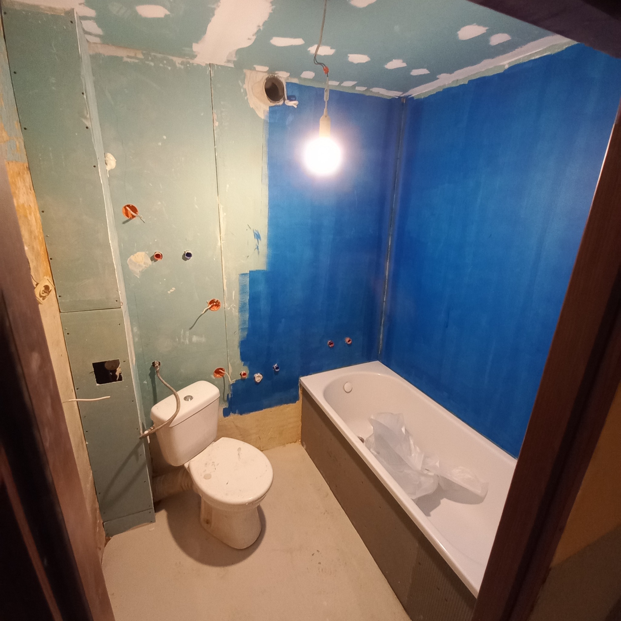 Łazienka w trakcie malowania: ściana pomalowana na niebiesko do połowy, widoczna wanna z folią ochronną, surowa ściana z instalacją elektryczną, wisząca żarówka.