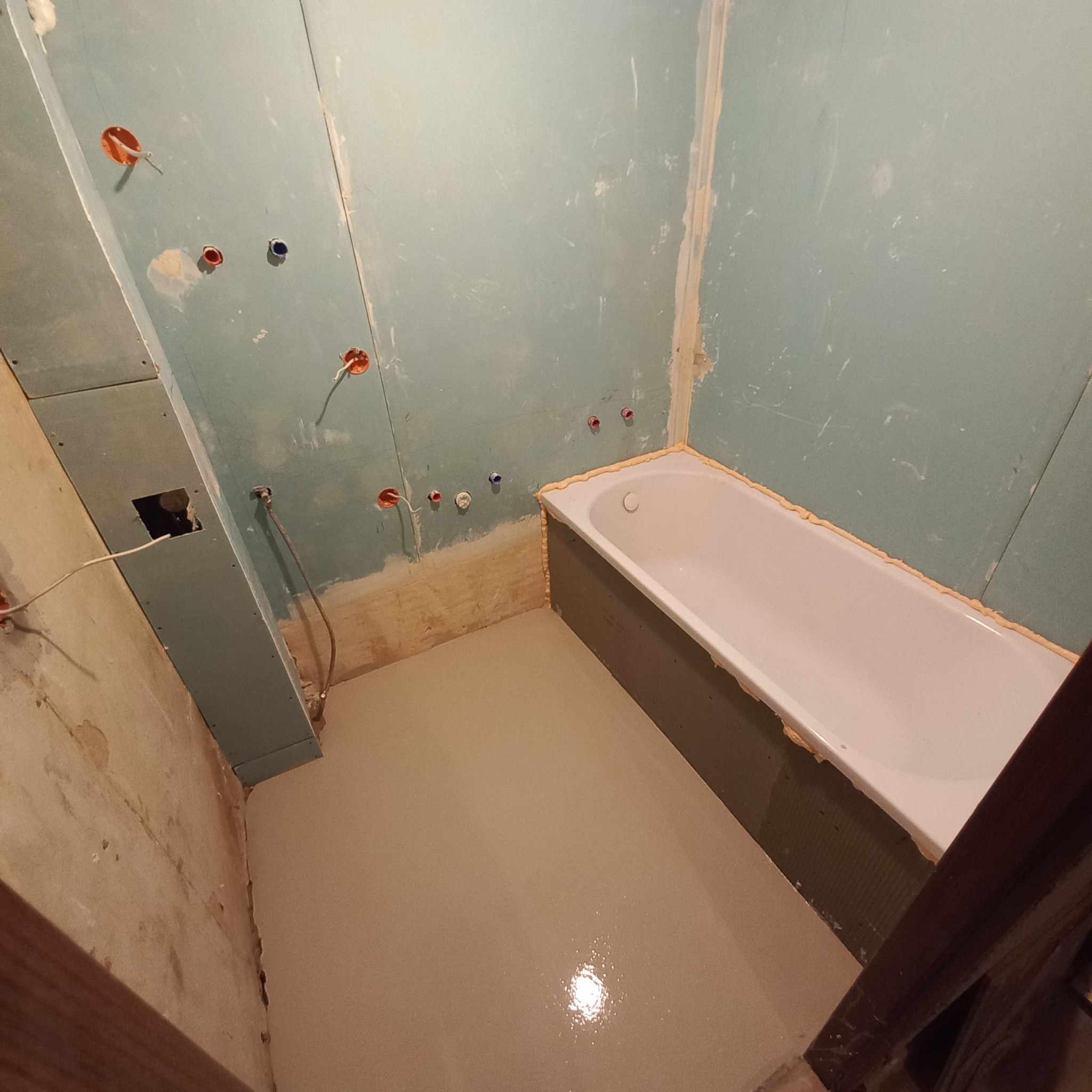 Łazienka w trakcie prac: wylewka samopoziomująca na podłodze, zamontowana wanna zabezpieczona pianką, ściany przygotowane pod płytki z widocznymi punktami instalacji elektrycznej i hydraulicznej.