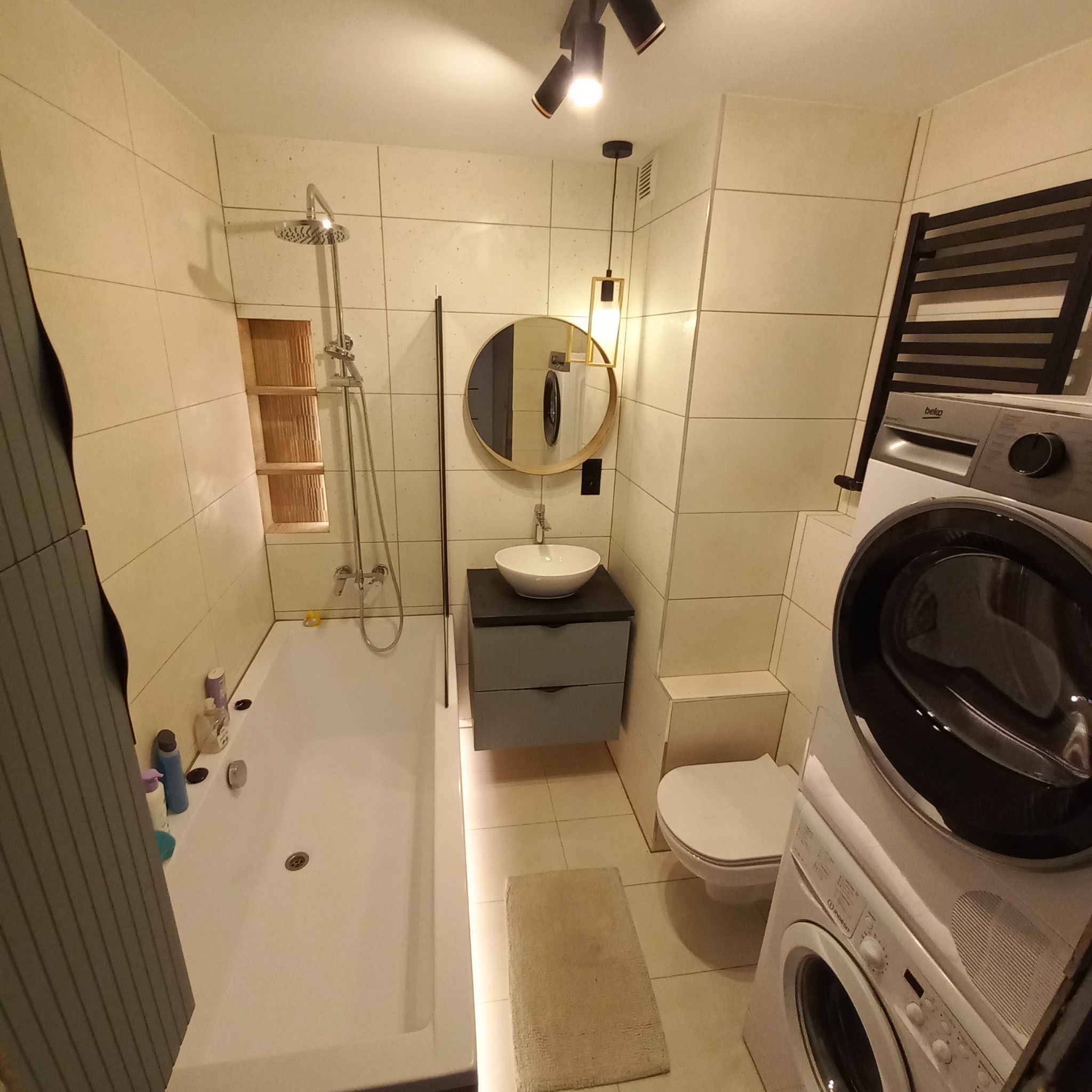 Jasna łazienka z wanną, umywalką na szafce, toaletą i pralką z suszarką, oświetlona punktowymi lampami i okrągłym lustrem.