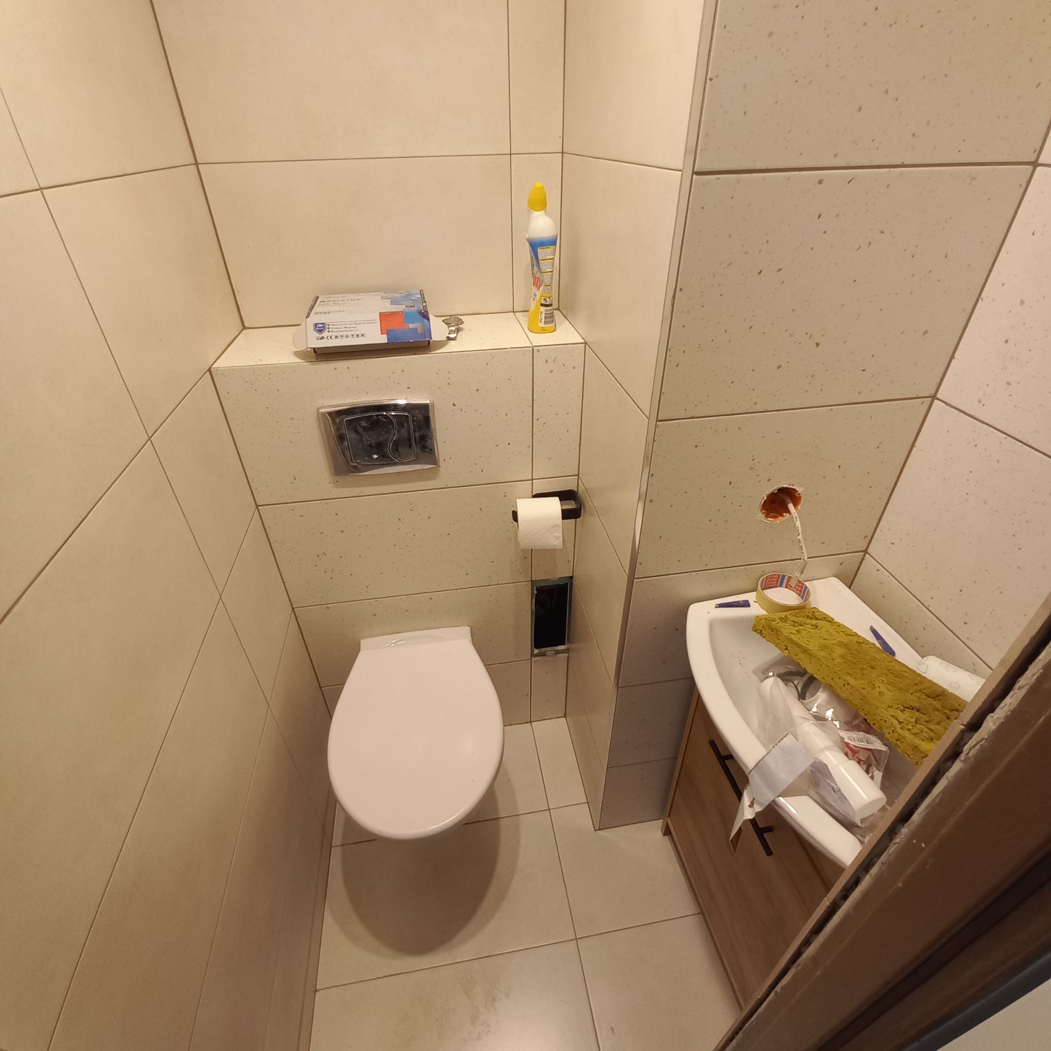 Niedokończona łazienka z wiszącą toaletą, umywalką z szafką, otwartym otworem na rurę i narzędziami w umywalce, płytki w odcieniach beżu.