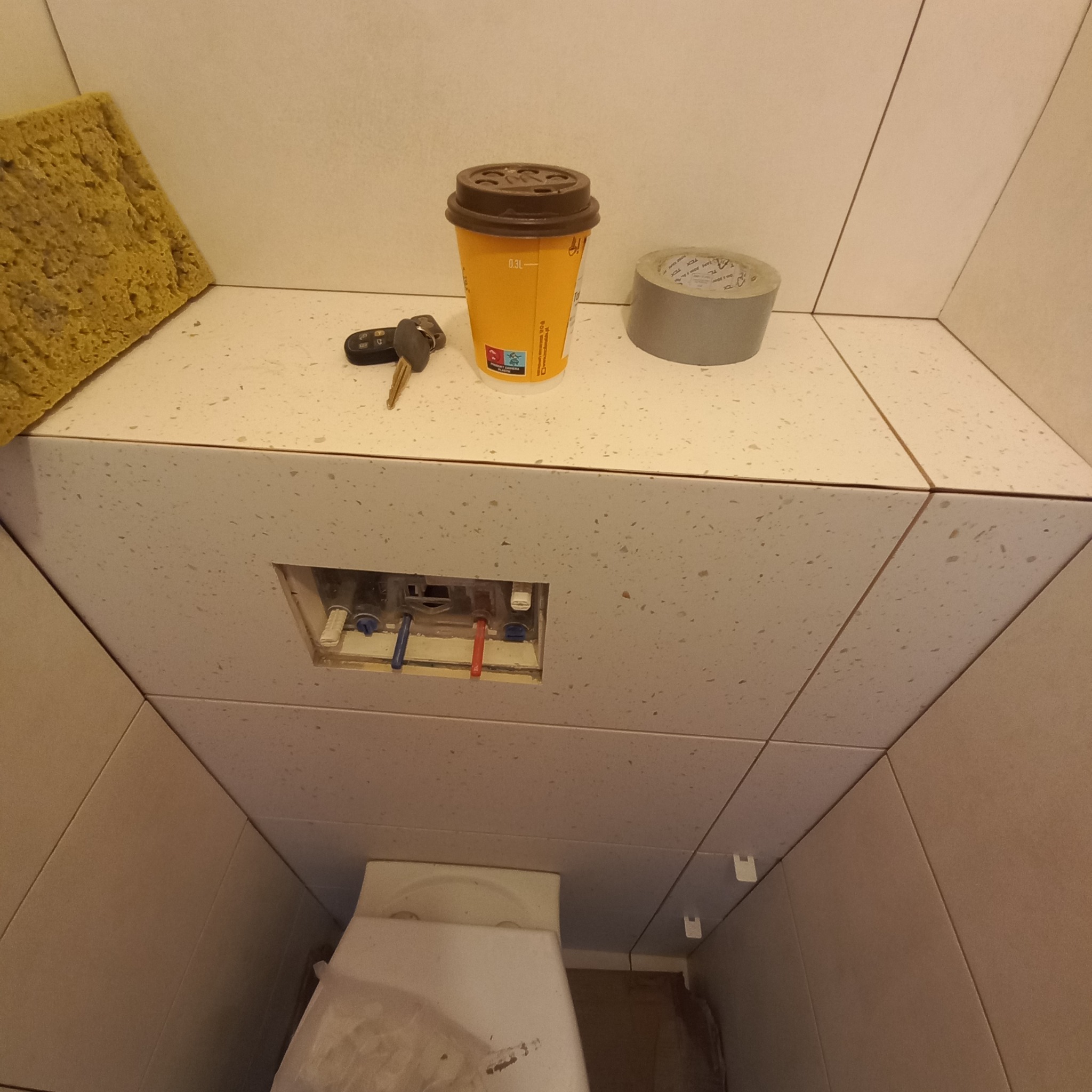 Niedokończona instalacja podtynkowa WC z widocznymi rurami, obok kubek po kawie, kluczyki samochodowe i taśma klejąca na blacie z płytek.