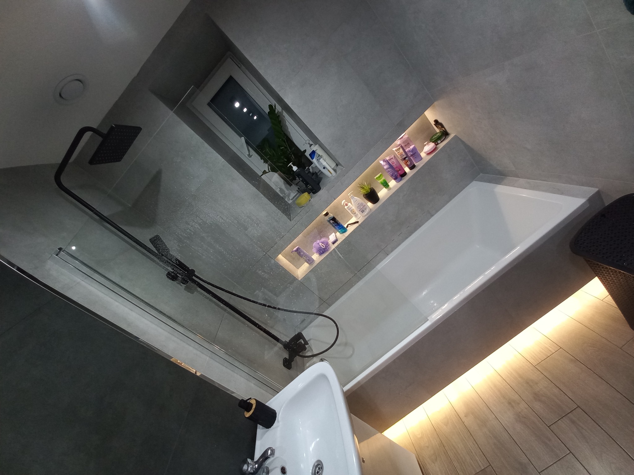 Nowoczesna łazienka z szarymi płytkami, czarną armaturą, podświetlaną wnęką na kosmetyki, oświetleniem pod wanną i rośliną w oknie.