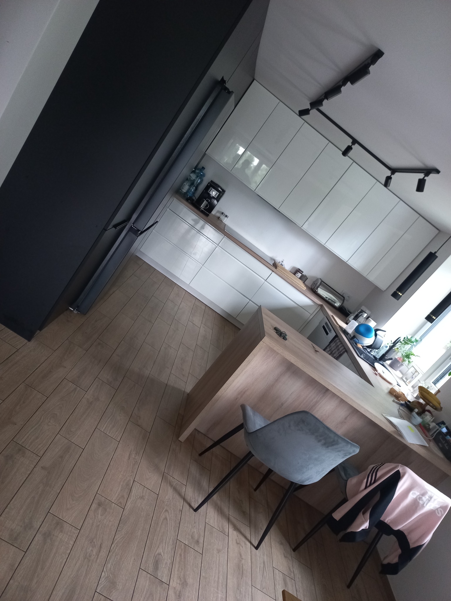 Nowoczesna kuchnia z białymi szafkami na wysoki połysk, drewnianym blatem, podłogą z paneli imitujących drewno i czarnymi akcentami oświetleniowymi.