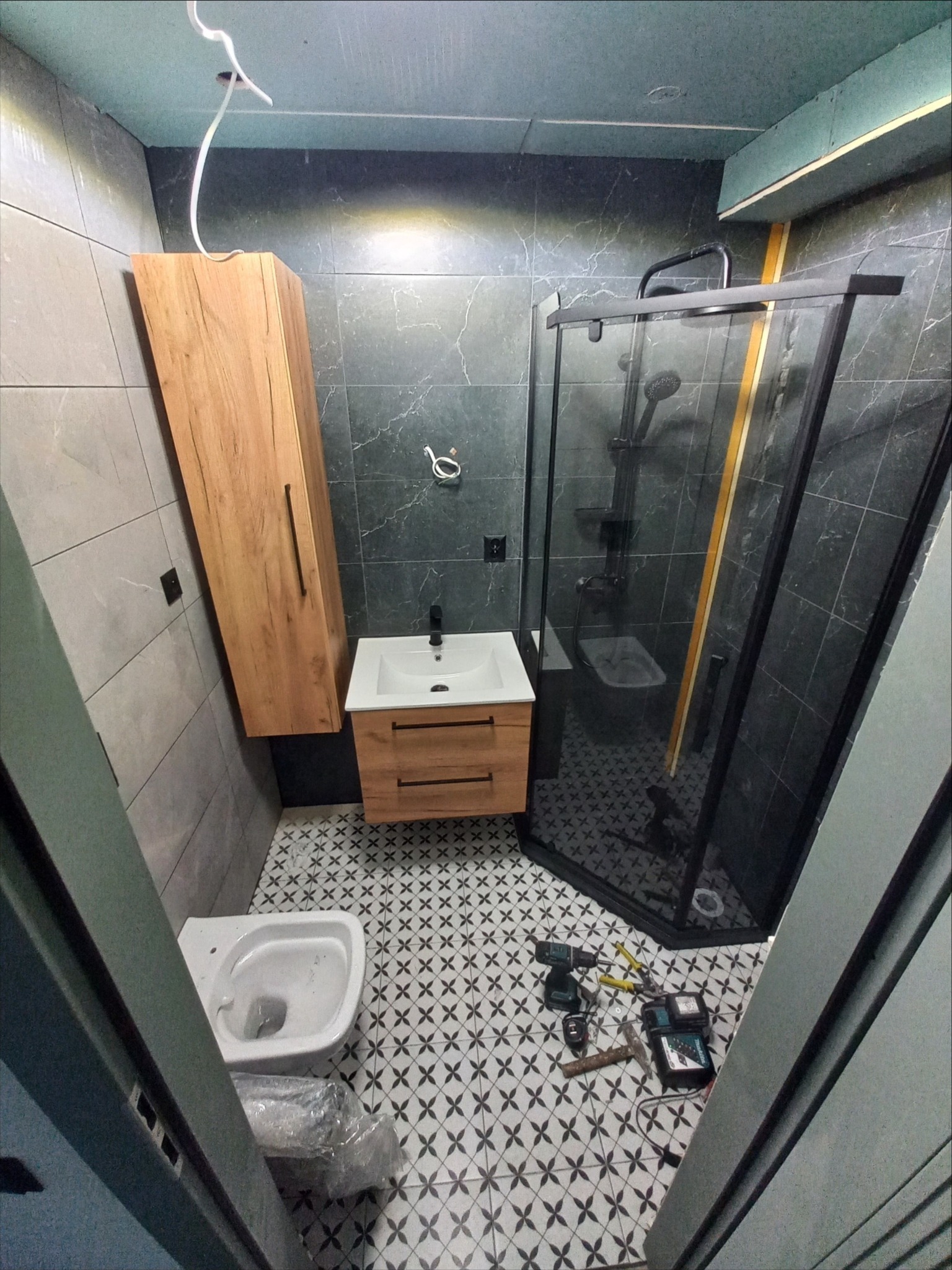 Wnętrze remontowanej łazienki w Piekarach Śląskich: kabina prysznicowa z czarnymi profilami, wisząca szafka z umywalką, toaleta w folii ochronnej i narzędzia glazurnicze na podłodze z wzorzystymi...