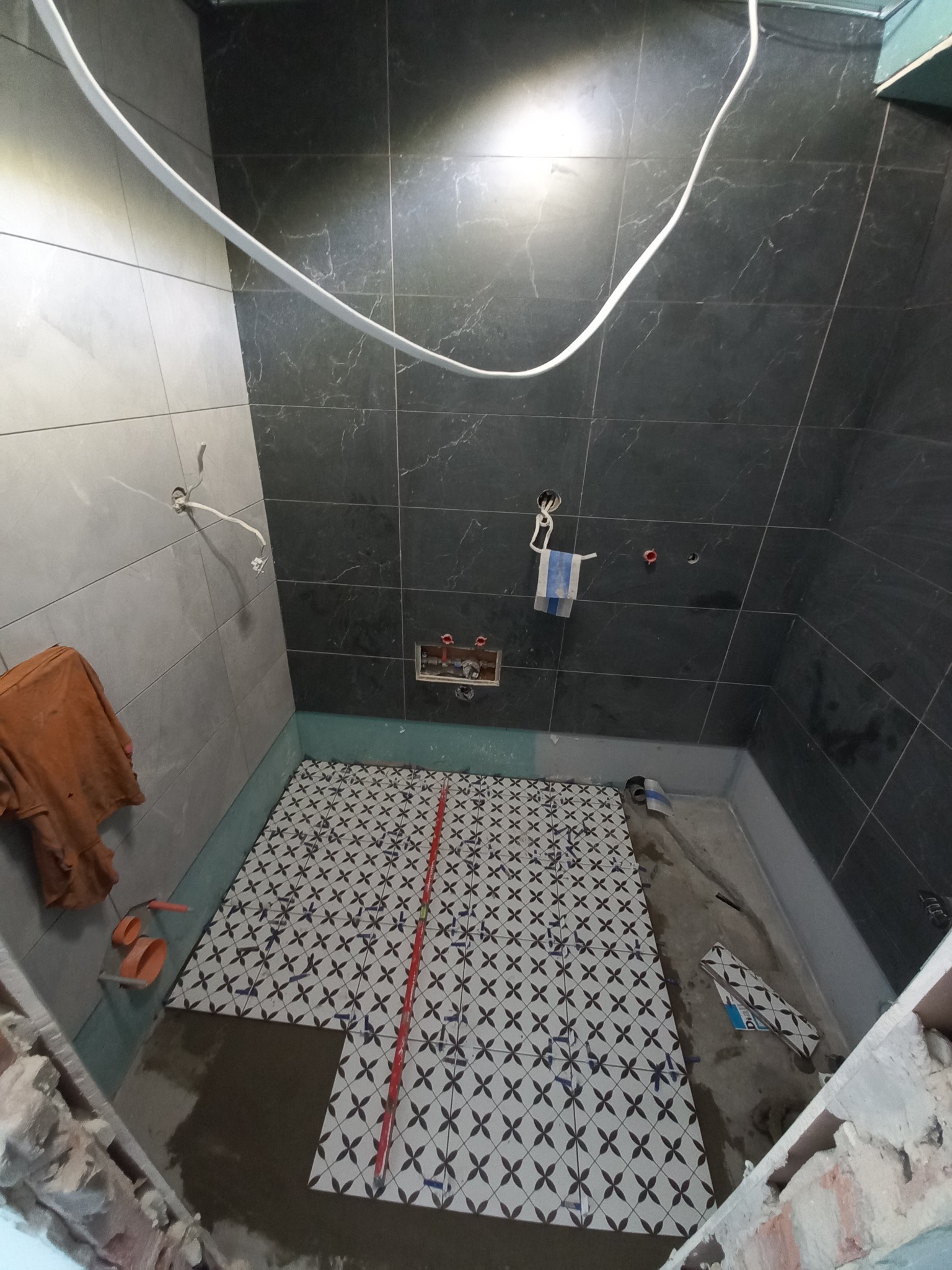 Montaż płytek podłogowych w łazience z wykorzystaniem wzorzystych płytek i czerwonego lasera krzyżowego, widoczne narzędzia glazurnicze i fragment ściany z szarymi płytkami.