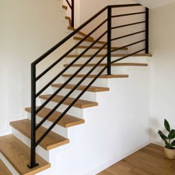 Metalowe Fantazje Radosław Panek-Porzelski - Nowoczesne schody z drewnianymi stopniami i czarną, metalową balustradą. Minimalistyczny design, jasne wnętrze, roślina doniczkowa w tle. Ujęcie z dołu, podkreślające geometrię.