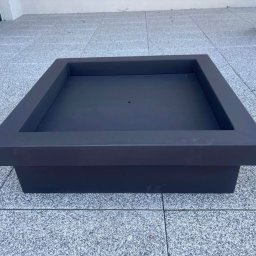 Metalowe Fantazje Radosław Panek-Porzelski - Kwadratowa, metalowa konstrukcja w kolorze grafitowym, prawdopodobnie donica lub obudowa paleniska, ustawiona na szarym, płytkowym podłożu. Minimalistyczny design.