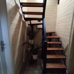 Metalowe Fantazje Radosław Panek-Porzelski - Drewniane schody z metalową konstrukcją w jasnym wnętrzu wykończonym boazerią. Obok schodów widoczne rośliny doniczkowe na małym stoliku.