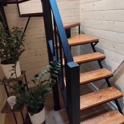 Metalowe Fantazje Radosław Panek-Porzelski - Drewniane schody z czarną metalową balustradą w jasnym wnętrzu z boazerią. Na podeście rośliny doniczkowe. Widok z dołu, ukazujący solidną konstrukcję i estetyczne wykończenie.
