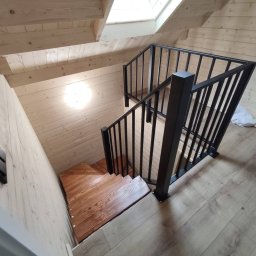 Metalowe Fantazje Radosław Panek-Porzelski - Drewniane schody z czarną, metalową balustradą w jasnym wnętrzu z boazerią. Widok z góry, podkreślający solidne wykonanie i nowoczesny design.