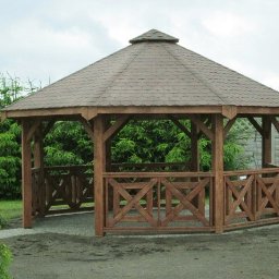 Metalowe Fantazje Radosław Panek-Porzelski - Drewniana altana ogrodowa z brązowym dachem, otoczona zielenią. Konstrukcja z krzyżowymi zdobieniami, widoczna na tle drzew i fragmentu ogrodzenia.