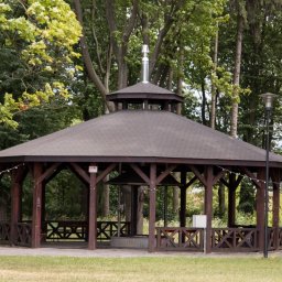 Metalowe Fantazje Radosław Panek-Porzelski - Drewniana altana ogrodowa z grillem pośrodku, otoczona zielenią. Konstrukcja z ciemnego drewna, zadaszona, z ozdobnymi barierkami. Widok z zewnątrz, w tle drzewa i trawnik.