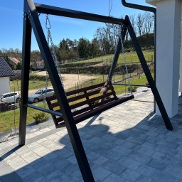 Metalowe Fantazje Radosław Panek-Porzelski - Metalowa huśtawka ogrodowa z drewnianym siedziskiem, zawieszona na łańcuchach, na tle kostki brukowej i zieleni. Konstrukcja pomalowana na czarno, widoczne detale mocowań.