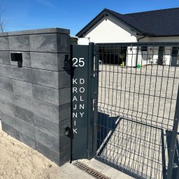 Metalowe Fantazje Radosław Panek-Porzelski - Nowoczesne ogrodzenie panelowe z szarego betonu i metalowej siatki, z numerem domu '25' i nazwą ulicy 'Królnik' na tle błękitnego nieba i domu.
