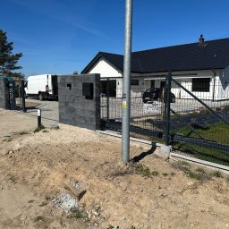 Metalowe Fantazje Radosław Panek-Porzelski - Nowoczesne ogrodzenie panelowe z bramą wjazdową na posesję z domem jednorodzinnym w słoneczny dzień. Widoczny fragment podjazdu i trawnika. Słupek oświetleniowy.