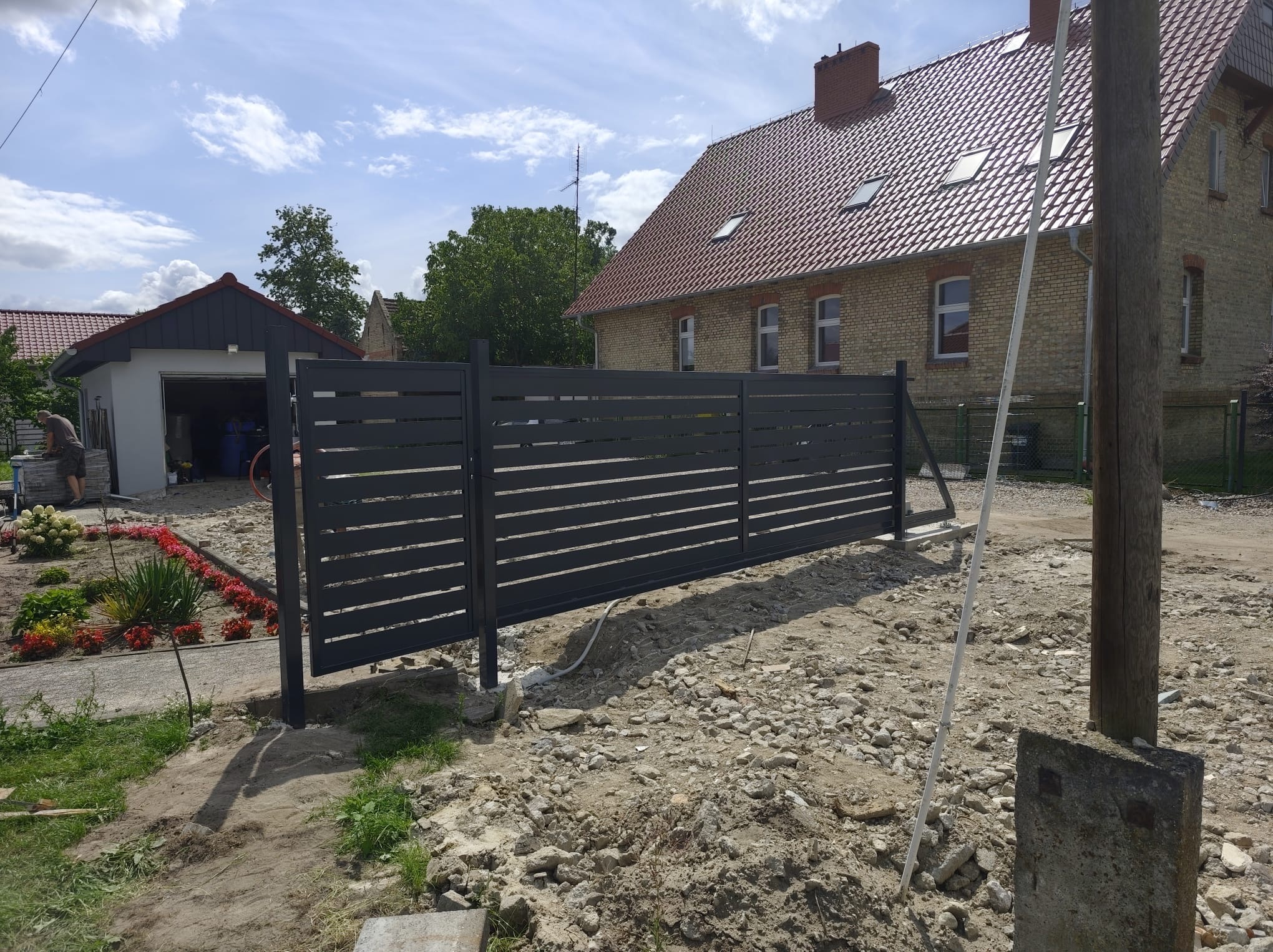 Nowoczesne, poziome przęsła ogrodzeniowe w kolorze grafitowym, w trakcie montażu na posesji z domem z czerwonej cegły. Widoczne elementy montażowe i przygotowany teren.